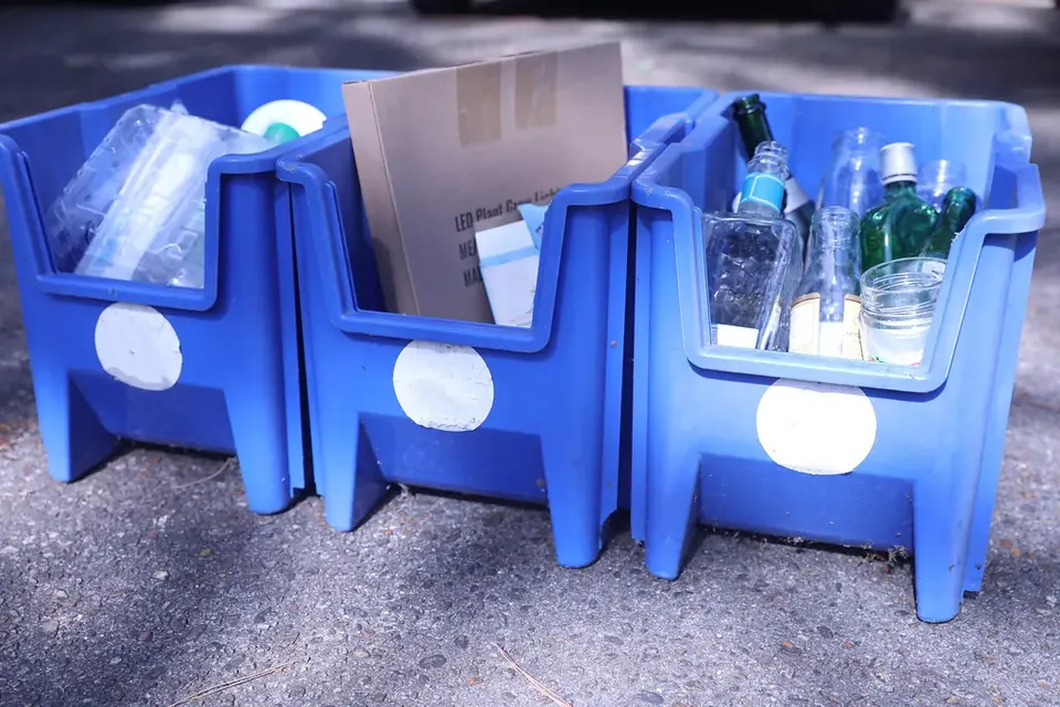 Three blue recycling bins outdoors, filled with glass bottles, plastic and cardboard